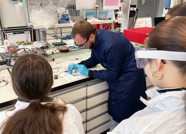 School students observing a researcher who is carrying out an experiment on electrochemistry.